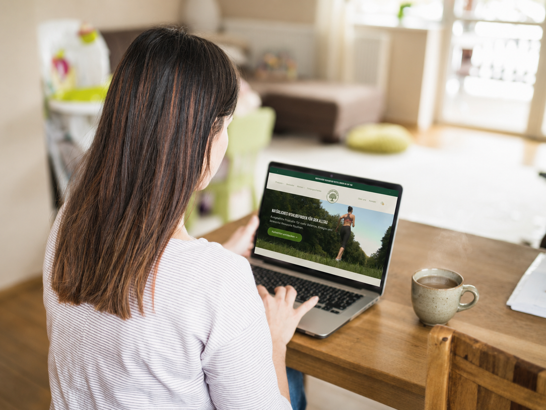 Frau nutzt die Reformwelt-Website auf ihrem Laptop und trinkt heissen Tee an einem Holztisch in einem hellen Zuhause.