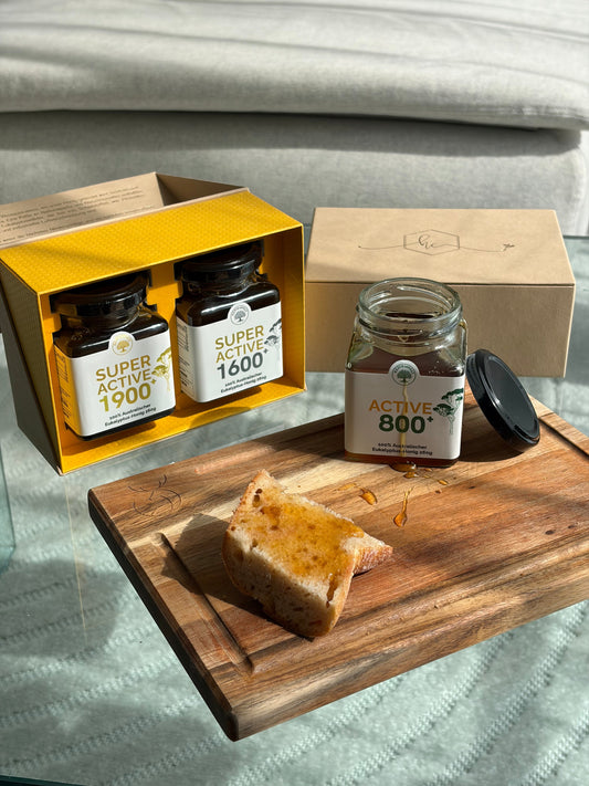 a gift box of bioactive on top of a table with a table cloth beside a chopping board with a bread drizzled with honey on top.
