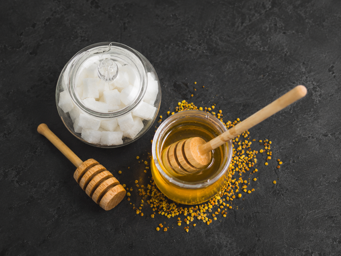 Jar of honey with honey dipper next to a glass jar of sugar cubes on a dark background.