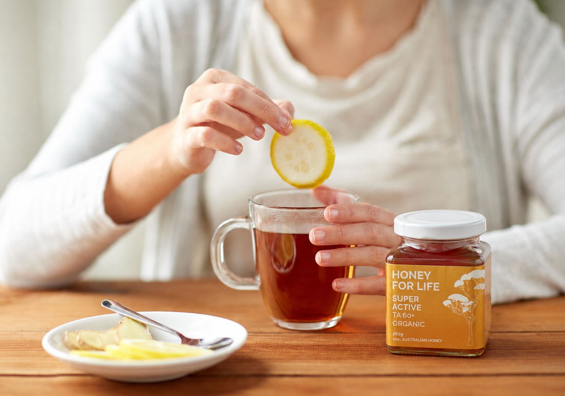 Person gibt Zitrone in eine Tasse Tee; daneben steht ein Glas bioaktiver Honig auf einem Holztisch.