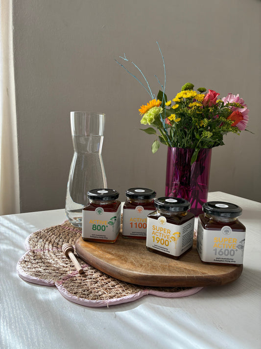 Four jars of Reformwelt Active and Super Active eucalyptus honey displayed on a wooden board beside a glass water carafe and colourful flowers.