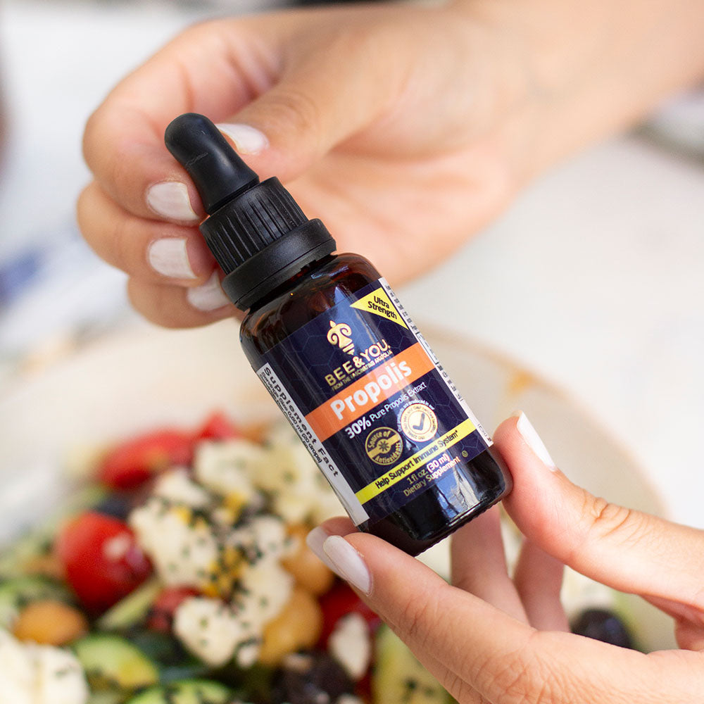 both hands holding a propolis bottle with a blurred background of a bowl of salad.